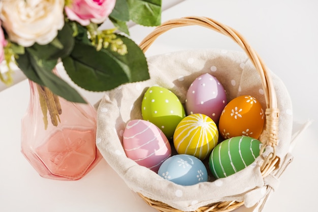 Huevos de Pascua coloridos en busket en mesa de madera blanca. Ramo de flores color de rosa en la mesa. Felices Pascuas. Estado de ánimo primaveral.