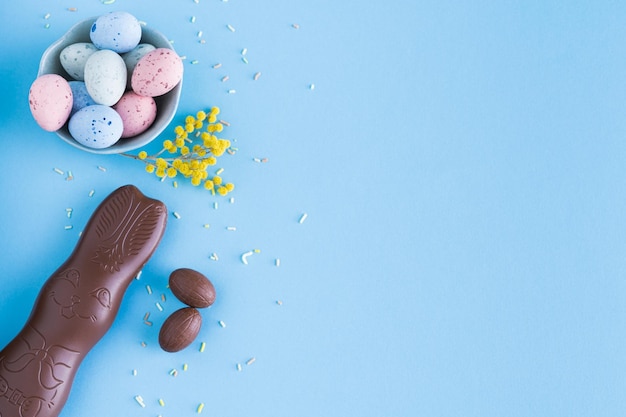 Huevos de Pascua y conejito de chocolate sobre fondo azul con decoraciones de flores amarillas Vista superior Espacio de copia