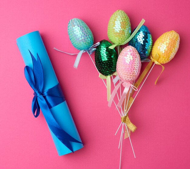 Huevos de Pascua decorativos multicolores decorados con lentejuelas y rollo de papel azul sobre un fondo rosa