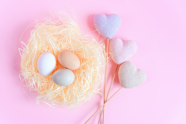 Huevos de Pascua de diferentes colores en un nido de paja y corazones textiles decorativos sobre una superficie rosa, vista superior, endecha plana. Concepto de pascua.