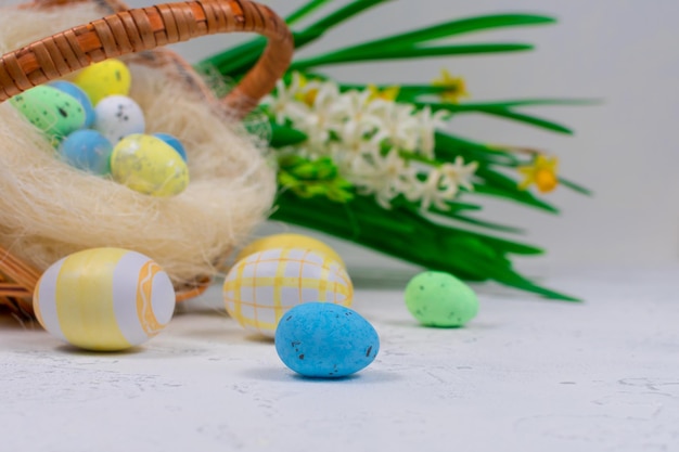 Huevos de Pascua multicolores en colores pastel sobre una mesa blanca con el telón de fondo de una cesta con huevos de Pascua y flores de narcisos Enfoque selectivo Primer plano