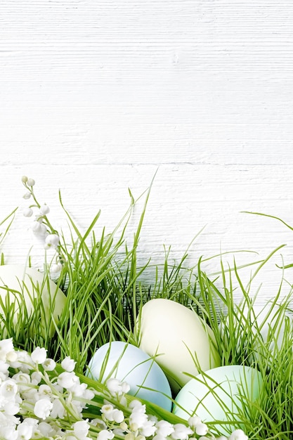 Huevos de Pascua en pasto verde con flores de lirios del valle. Decoraciones de vacaciones de Pascua sobre fondo blanco de madera.