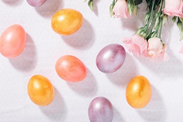 Huevos de Pascua pintados de púrpura y rosa dorados en colores pastel y flores sobre un mantel de algodón blanco