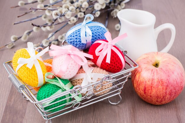 Huevos de Pascua tejidos atados con cintas de colores en una canasta de metal, una manzana, una jarra y sauce sobre una mesa de madera