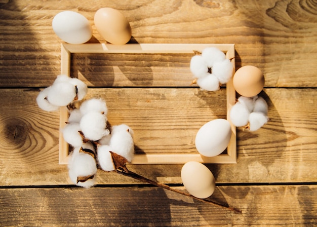 Huevos y una rama con flores de algodón blanco con sombras soleadas sobre un fondo de madera. Las ramitas se entrelazan con un marco de madera. Una tarjeta de vacaciones. El concepto de la fiesta de Pascua.