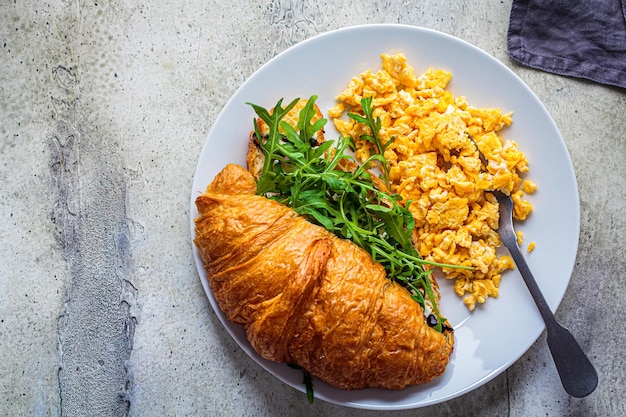Huevos revueltos y croissant con queso y rúcula en la vista superior de un plato blanco Huevos y croissant para el desayuno Concepto de desayuno