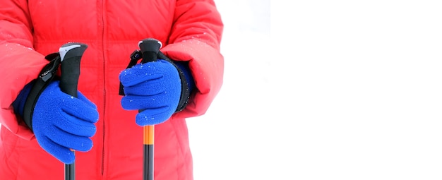 Humano em uma jaqueta esportiva vermelha e luvas azuis segura duas varas para caminhadas no inverno, isoladas no fundo branco, com espaço livre para texto no lado direito da foto