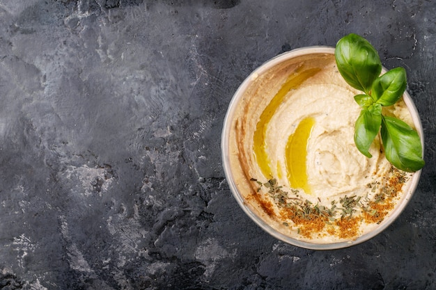 Hummus con aceite de oliva, pan de pita y comino molido en cuenco de cerámica servido con limones, albahaca y garbanzos sobre una superficie de textura oscura. Vista superior, endecha plana