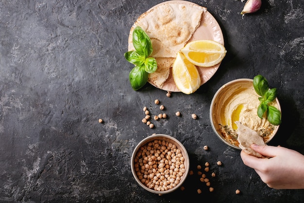 Hummus con aceite de oliva, pan de pita y comino molido en cuenco de cerámica servido con limones, albahaca y garbanzos sobre una superficie de textura oscura. Vista superior, endecha plana