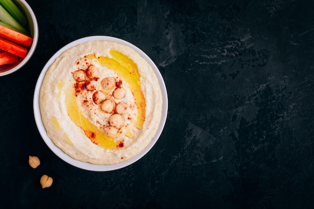 Hummus com azeite e grão de bico em tigela com palitos de vegetais no fundo escuro de pedra Vista superior com espaço de cópia