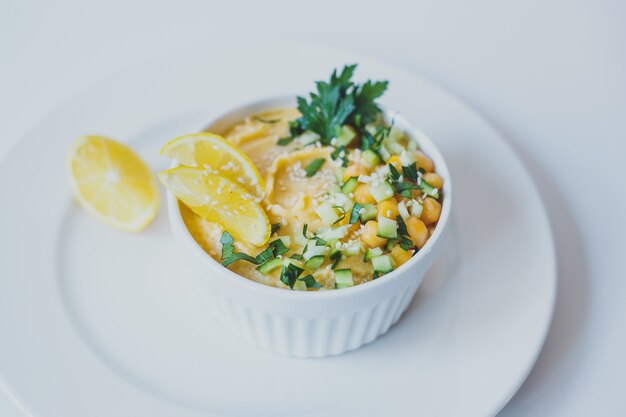 Hummus de garbanzos con aceite de oliva limón semillas de sésamo y pepinos sobre fondo blanco.