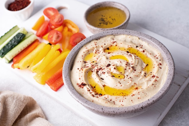 Hummus de garbanzos en un recipiente con rodajas de verduras en la placa de corte salsa vegana sobre fondo blanco.