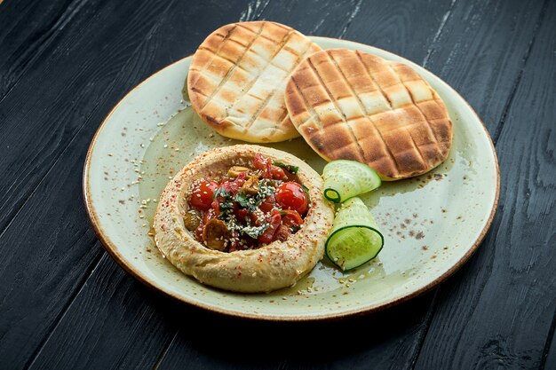 Hummus de garbanzos con tomate al horno y aceite de oliva servido con pita al horno en un plato sobre un fondo de madera oscura.