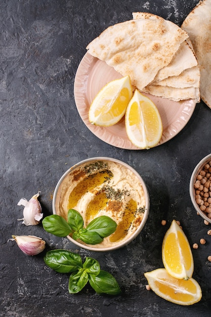 Hummus mit Olivenöl, Fladenbrot und gemahlenem Kreuzkümmel in einer Keramikschale, serviert mit Zitronen, Basilikum und Kichererbsen über einer dunklen Texturoberfläche. Draufsicht, flach liegen