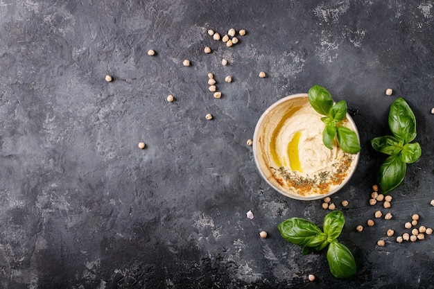 Hummus mit Olivenöl, Fladenbrot und gemahlenem Kreuzkümmel in einer Keramikschale, serviert mit Zitronen, Basilikum und Kichererbsen über einer dunklen Texturoberfläche. Draufsicht, flach liegen