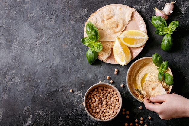 Hummus mit Olivenöl, Fladenbrot und gemahlenem Kreuzkümmel in einer Keramikschale, serviert mit Zitronen, Basilikum und Kichererbsen über einer dunklen Texturoberfläche. Draufsicht, flach liegen