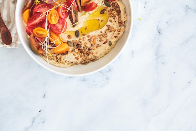 Hummus con plántulas de tomate, nueces y aceite de oliva en un plato blanco con fondo de mármol blanco