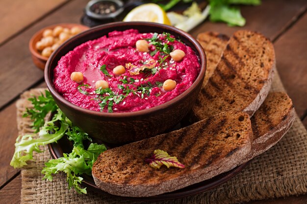 Hummus de remolacha asado con tostadas en un tazón de cerámica sobre una mesa de madera.