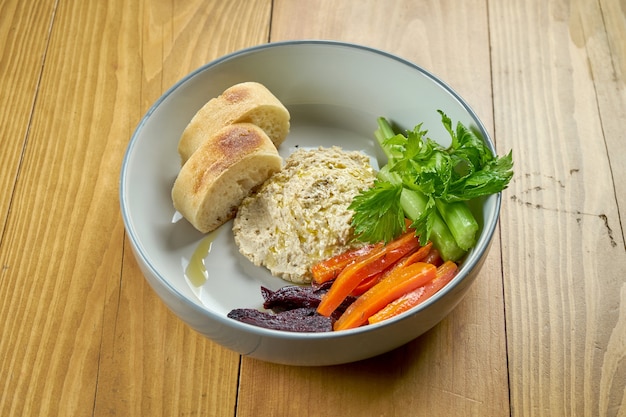 Hummus con verduras al horno y baguette en un recipiente sobre un fondo de madera. Plato vegetariano. Cocina israelí