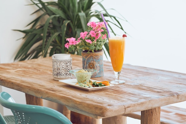 Hummus y verduras cortadas servidas en un plato blanco cerca de un vaso de jugo y flores en una maceta sobre una mesa de madera en un comedor luminoso