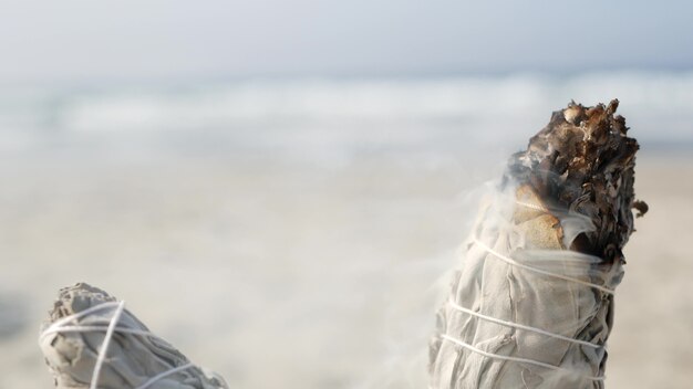 Humo de salvia blanca seca, quema de palitos de manchas secas, bokeh de enfoque suave. Manchas de aroma de cerca en la playa del océano, meditación de aromaterapia por las olas del agua de mar en la costa arenosa. Yoga, paz, armonía y ayurveda