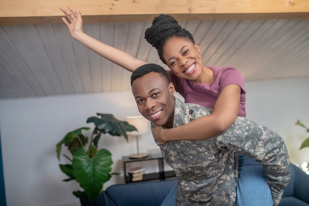 Humor divertido. Joven militar afroamericano sosteniendo esposa gesticulando hacia atrás en estado de ánimo lúdico en casa en habitación acogedora