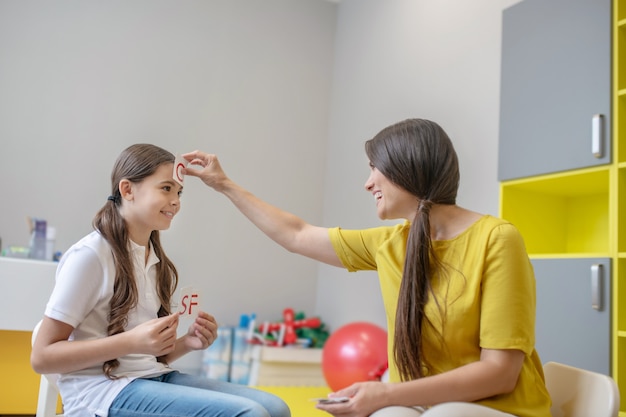 Foto humor divertido. psicóloga sonriente alegre que realiza terapia de juego con una chica alegre y sociable usando tarjetas de letras