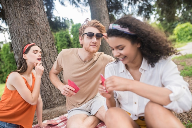 Foto humor divertido. tres jóvenes amigos felices divirtiéndose jugando a las cartas sentados en una manta al aire libre en verano