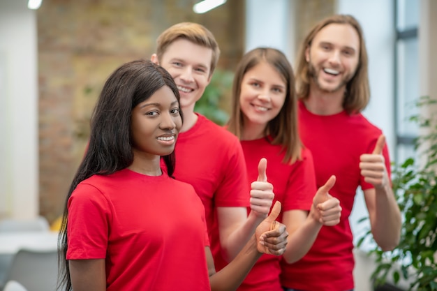 Humor otimista. jovem americana e amigos caucasianos se voluntários em camisetas correspondentes em pé juntos, mostrando um gesto de ok