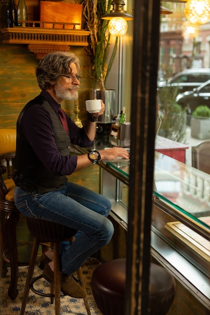 Humor pensativo. Homem bonito pensativo sentado à mesa do café, bebendo café e olhando pela janela para as pessoas que passavam.