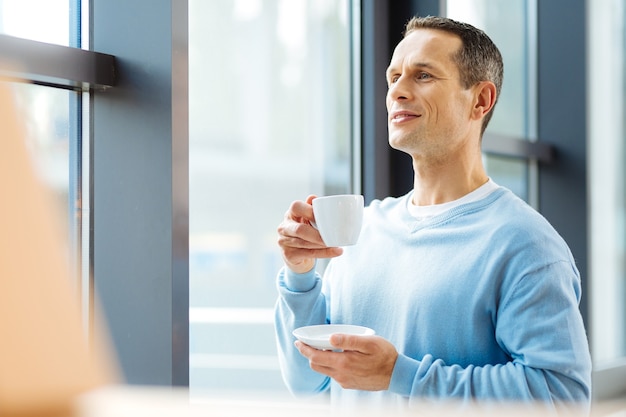 Humor positivo. Agradable hombre agradable alegre mirando por la ventana y sonriendo mientras toma un café