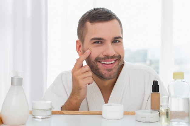 Humor positivo. homem simpático e encantado tocando seu rosto enquanto aplica o creme facial