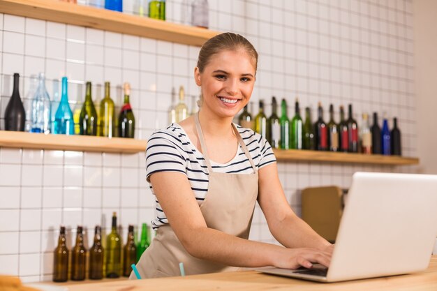 Humor positivo. Mulher simpática e alegre usando avental e sorrindo para você enquanto trabalha no laptop