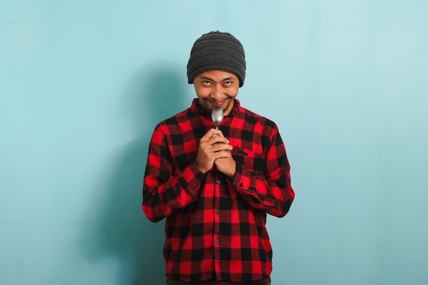 Humoroso Jovem asiático segurando colher perto de sua boca sorrindo maliciosamente isolado em fundo azul