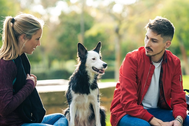 Hund mit Aufmerksamkeitsausdruck sitzt zwischen einem jungen Mann und einer Frau in einem öffentlichen Park