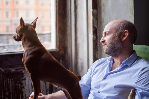 Hund und sein Besitzer schauen nachdenklich aus dem Fenster und beobachten die Straße Freundschaft zwischen einem Hund und einem Mann