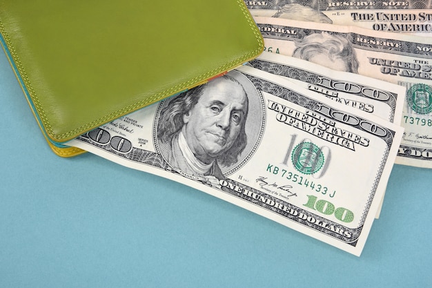 Hundert-Dollar-Scheine spähen aus einer grünen Lederbrieftasche auf einem türkisfarbenen Hintergrund.