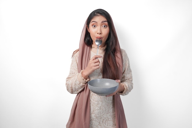 Hungrige asiatische muslimische Frau hält eine Schüssel und denkt darüber nach, was sie essen soll, während sie einen Löffel über der Schüssel beißt und auf den Iftar wartet. Isolierter weißer Hintergrund