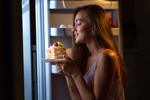 Hungrige frau im schlafanzug, die süßen kuchen nachts nahe kühlschrank isst