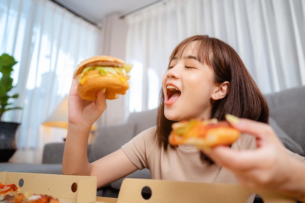 Hungrige junge Frau, die Junk-Food-Hamburger und Pizza zum Mittagessen isst, indem sie im Urlaub die Lieferung zu Hause bestellt. ungesunde Mahlzeit, Fettleibigkeitsrisiko.