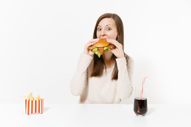 Hungrige junge Frau isst gierig Burger, sitzt am Tisch mit Pommes Frites, Cola in der Flasche isoliert auf weißem Hintergrund. Richtige Ernährung oder amerikanisches klassisches Fast Food. Werbefläche mit Kopienraum.
