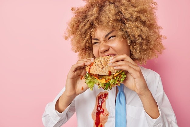 Hungrige junge frau mit lockigem haar beißt appetitlichen hamburger isst gerne fast food trägt weißes hemd mit ketchup und krawatte isoliert über rosa hintergrund ungesundes ernährungskonzept