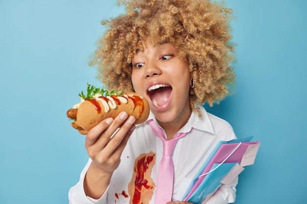 Hungrige Studentin isst leckeren Hotdog mit Grillwurst hat leckeren Snack genießt Fast Food hat keine Zeit für die Zubereitung von Mahlzeiten bereitet sich auf Prüfungen vor hält Ordner trägt weißes Hemd, das mit Ketchup verdreckt ist