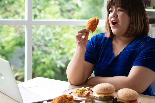 Hungrige übergewichtige Frau mit Fried Chicken Hamburger auf einer Holzplatte und Pizza auf dem Tisch