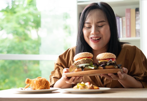 Hungrige übergewichtige Frau mit Hamburger auf einer Holzplatte Fried Chicken und Pizza auf dem Tisch