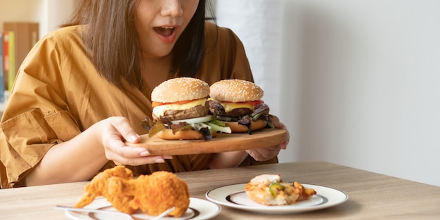 Hungrige überladene Frau, die Hamburger auf hölzerner Platte, gebratenem Huhn und Pizza auf Tabelle hält