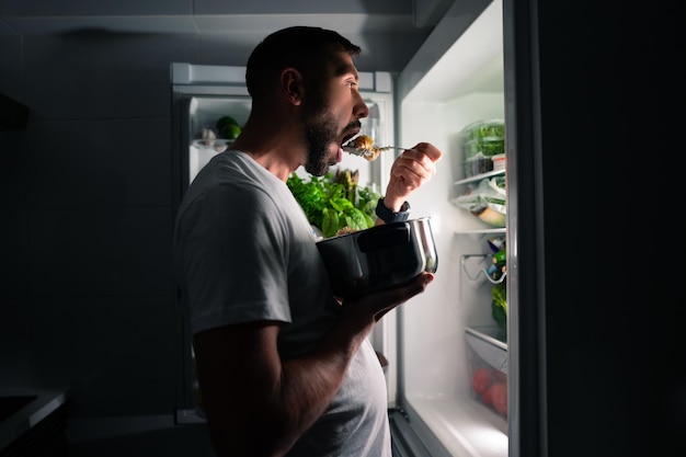 Hungriger Mann, der nachts Essen aus dem offenen Kühlschrank isst Mann, der Mitternachtssnack aus dem Kühlschrank nimmt