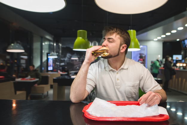 hungriger Mann isst einen großen appetitlichen Burger an einem Tisch in einem Fast-Food-Restaurant. Fast-Food-Konzept