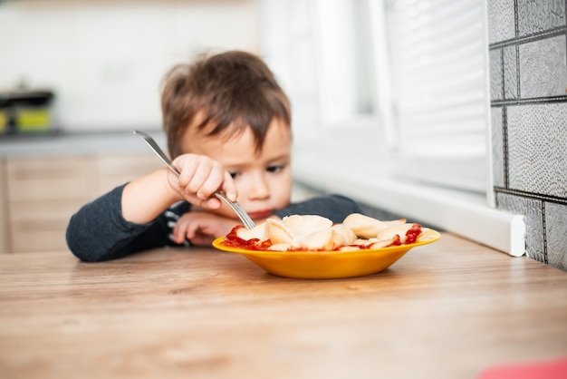 Hungriges Kind, das in der Küche Knödel isst und in einer grauen Jacke am Tisch sitzt