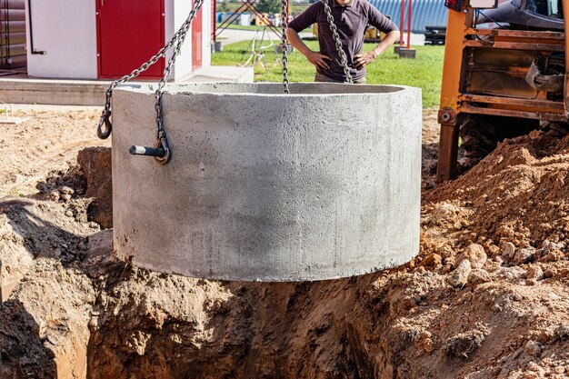 Hydraulisches Kolbensystem des Baggers mit einer Schaufel, das an Stahlseilen Betonkanalring in die Grube absenkt. Bau oder Reparatur eines Kanalhauses.